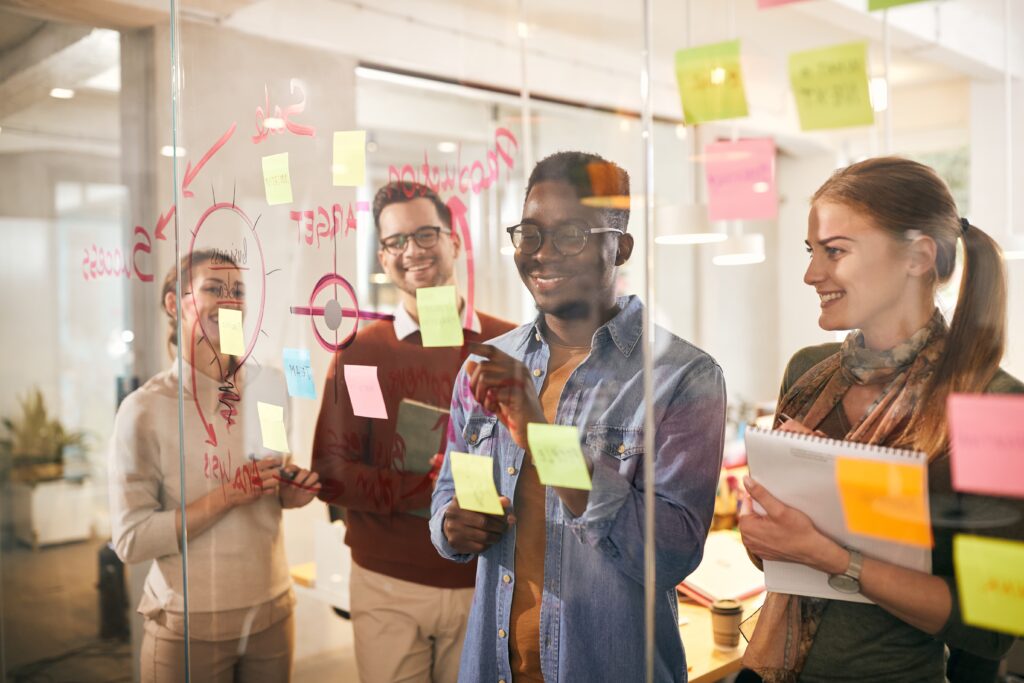 Équipe diverse devant un tableau en verre couvert de post-its colorés et de schémas pour planifier leur stratégie de communication