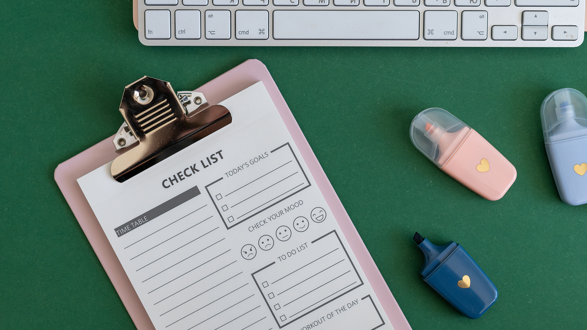 Presse-papiers avec une checklist posé sur un bureau vert, entouré d'un clavier et de surligneurs colorés.