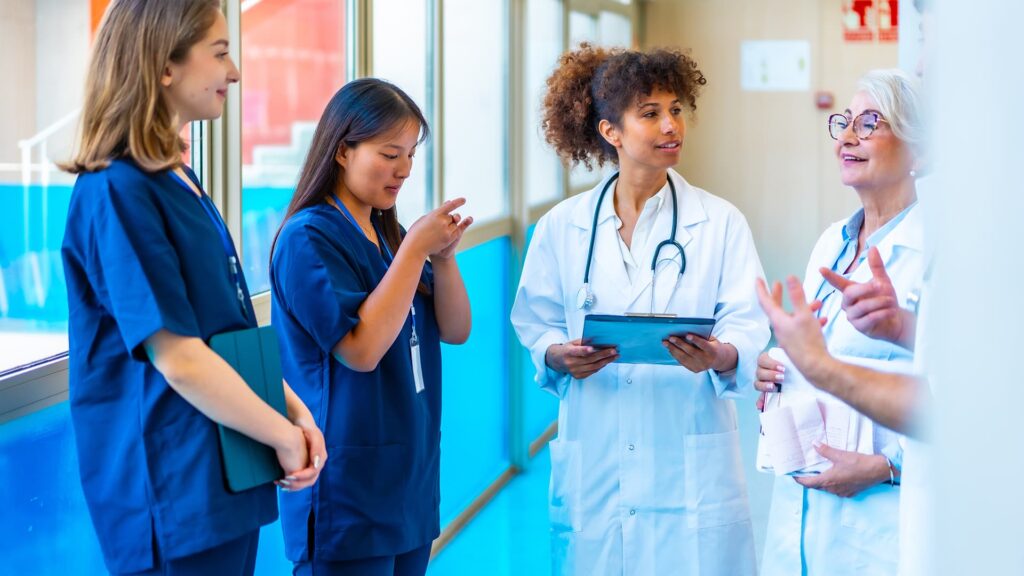 Quatre professionnelles de santé, deux infirmières en tenue bleue et deux médecins en blouse blanche, échangent dans un couloir d'hôpital, l'une tenant une tablette numérique.