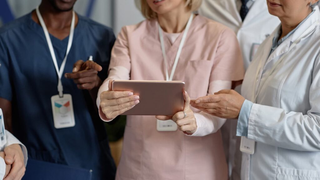 Plusieurs professionnels de santé aux tenues différentes, soignants et médecin en blouse blanche, consultent ensemble une tablette numérique lors d'un échange de coordination.