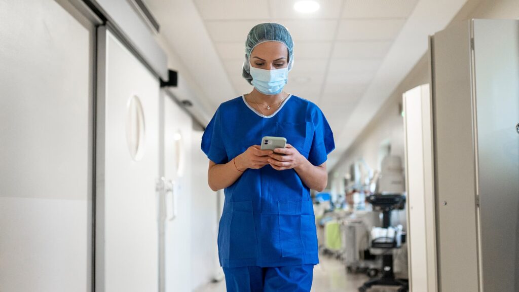 Une soignante en tenue bleue et masque chirurgical consulte son smartphone en marchant dans un couloir d'hôpital.