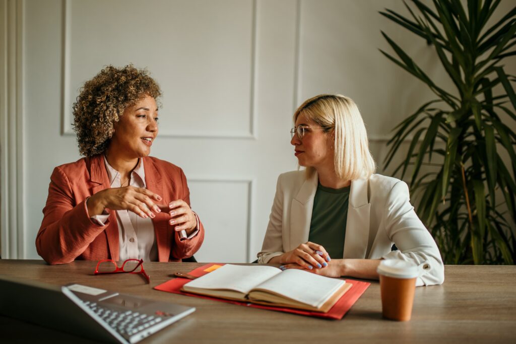 Deux collaboratrices en conversation lors d'un entretien professionnel, assises face à face autour d'une table avec un carnet ouvert et un ordinateur.