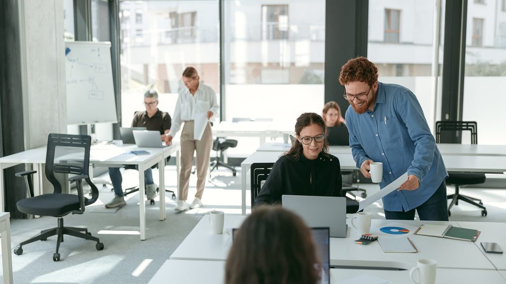 Un collaborateur aide une collègue devant son ordinateur portable dans un open space, pendant que d’autres travaillent en autonomie