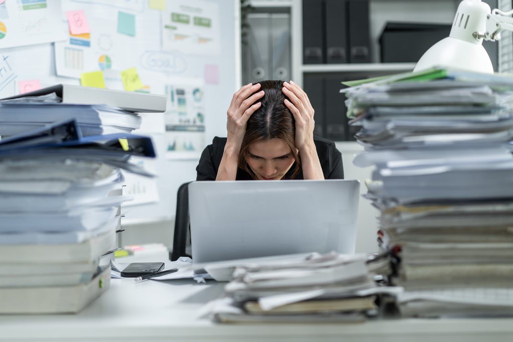 Salariée débordée, la tête entre les mains, entourée de piles de dossiers.